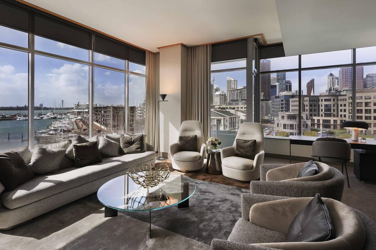 A modern living room with a sofa, armchairs, a glass coffee table, and a view of a marina and cityscape.