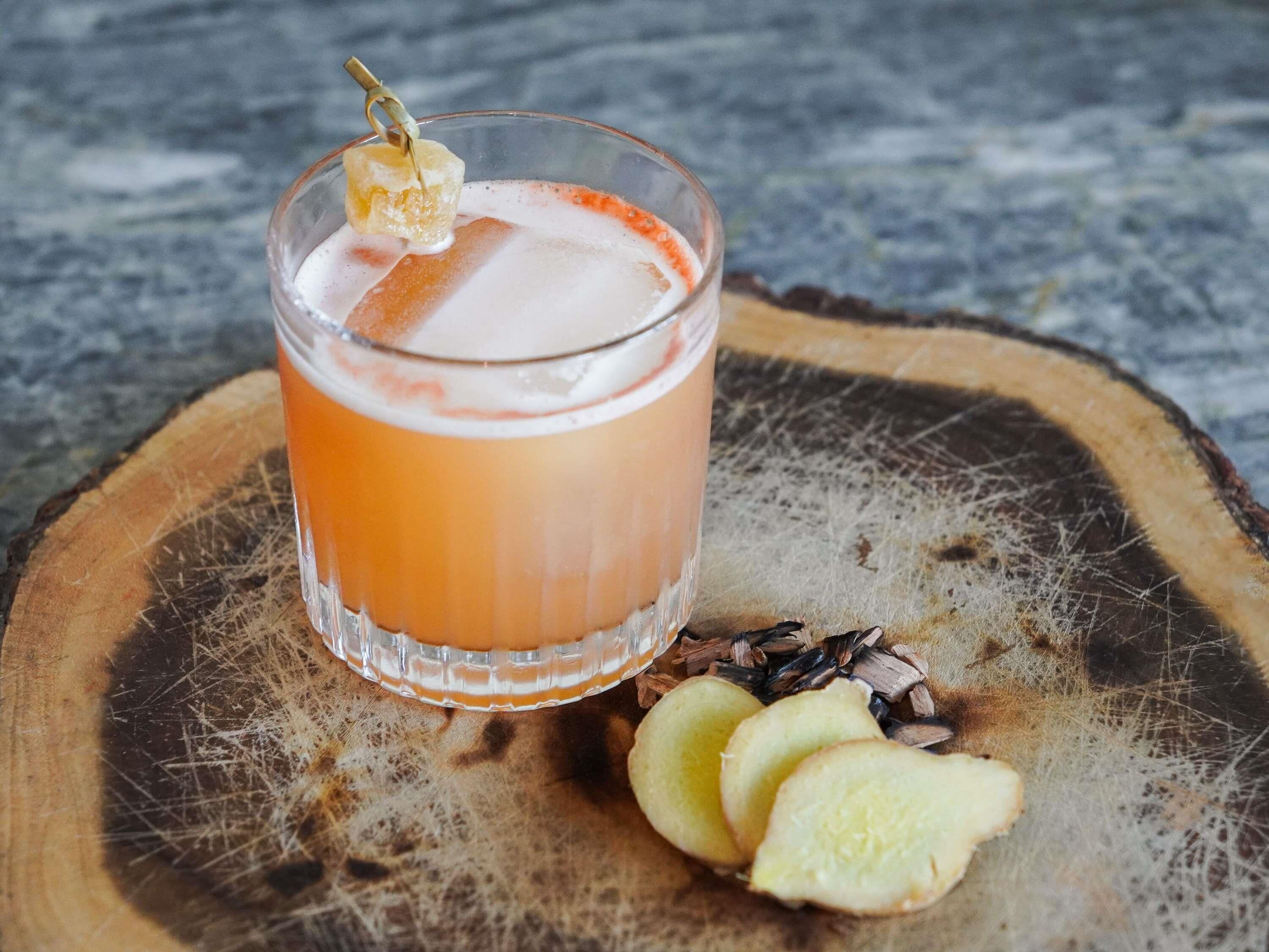 A glass of orange beverage with a garnish, placed on a wooden surface with scattered seeds and apple slices.