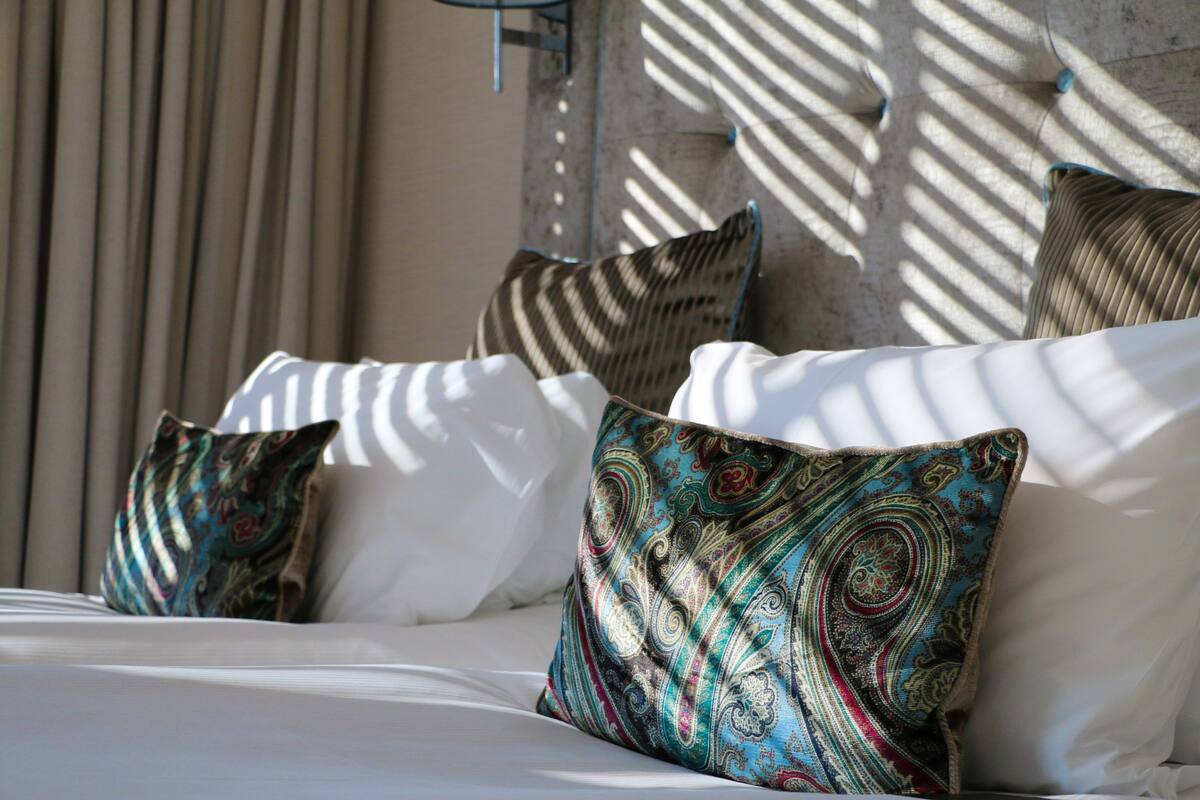 A bed with white linens and patterned pillows, a wall with a textured surface, and a window casting shadows.