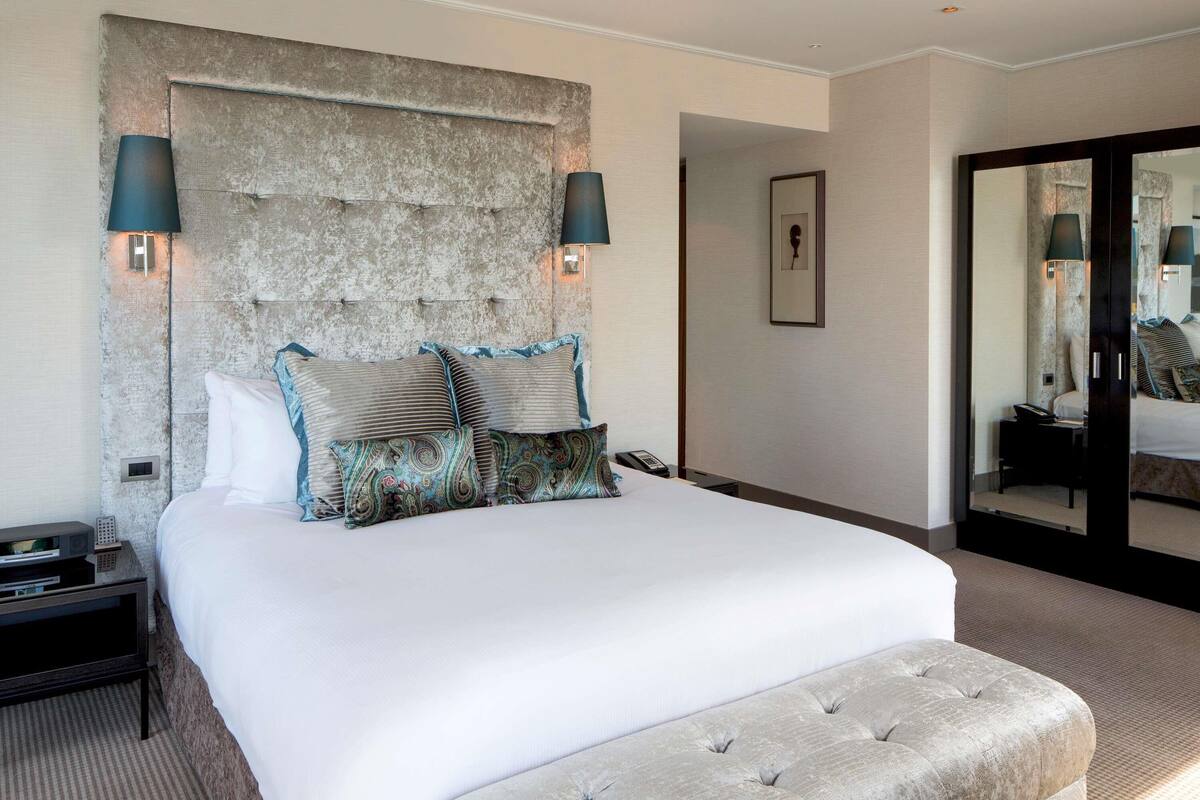 A hotel room with a large bed, a headboard with a textured stone-like design, two bedside lamps, and a large mirror on the wall.