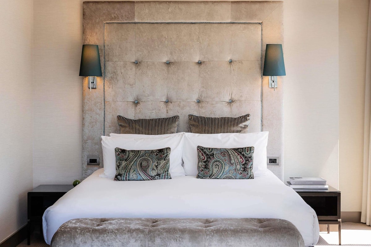 A modern bedroom with a tufted headboard, two bedside lamps, a bed with white linens, patterned pillows, and a small bedside table.