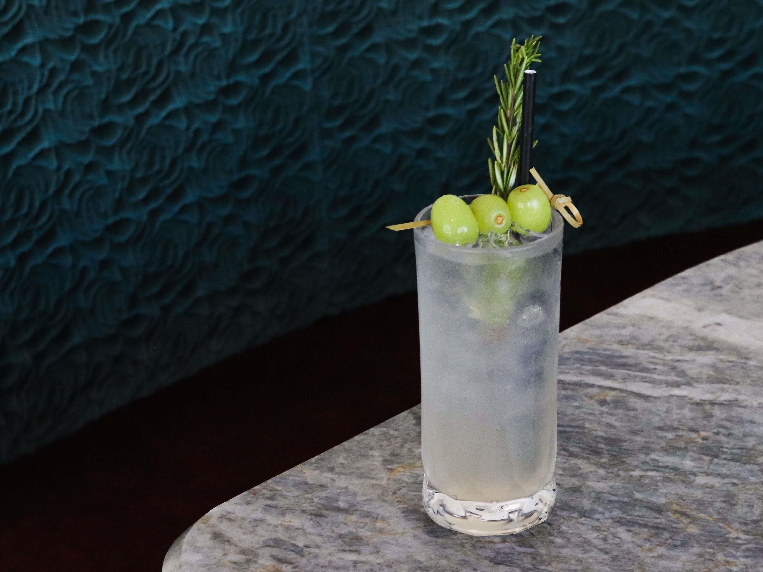A tall glass with a clear liquid, garnished with green olives, a sprig of rosemary, and a straw.