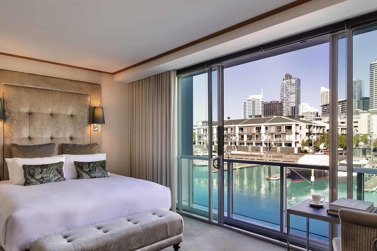 Habitación de hotel con una cama grande, cabecero capitonado y vistas a un puerto deportivo y el horizonte de la ciudad.