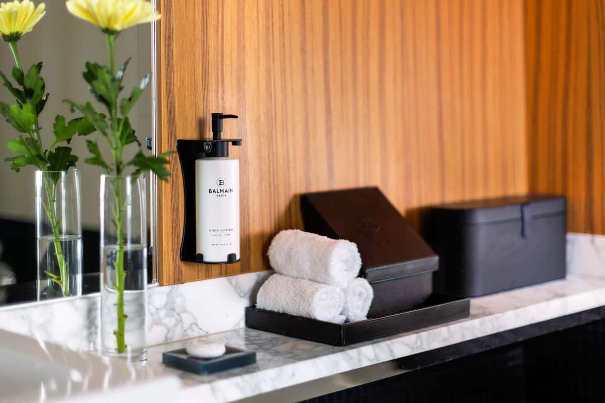 Un meuble-lavabo de salle de bain avec un plan de toilette en marbre, un flacon de lotion pour le corps de la marque Balmain, une trousse de toilette noire et un vase avec des fleurs jaunes.
