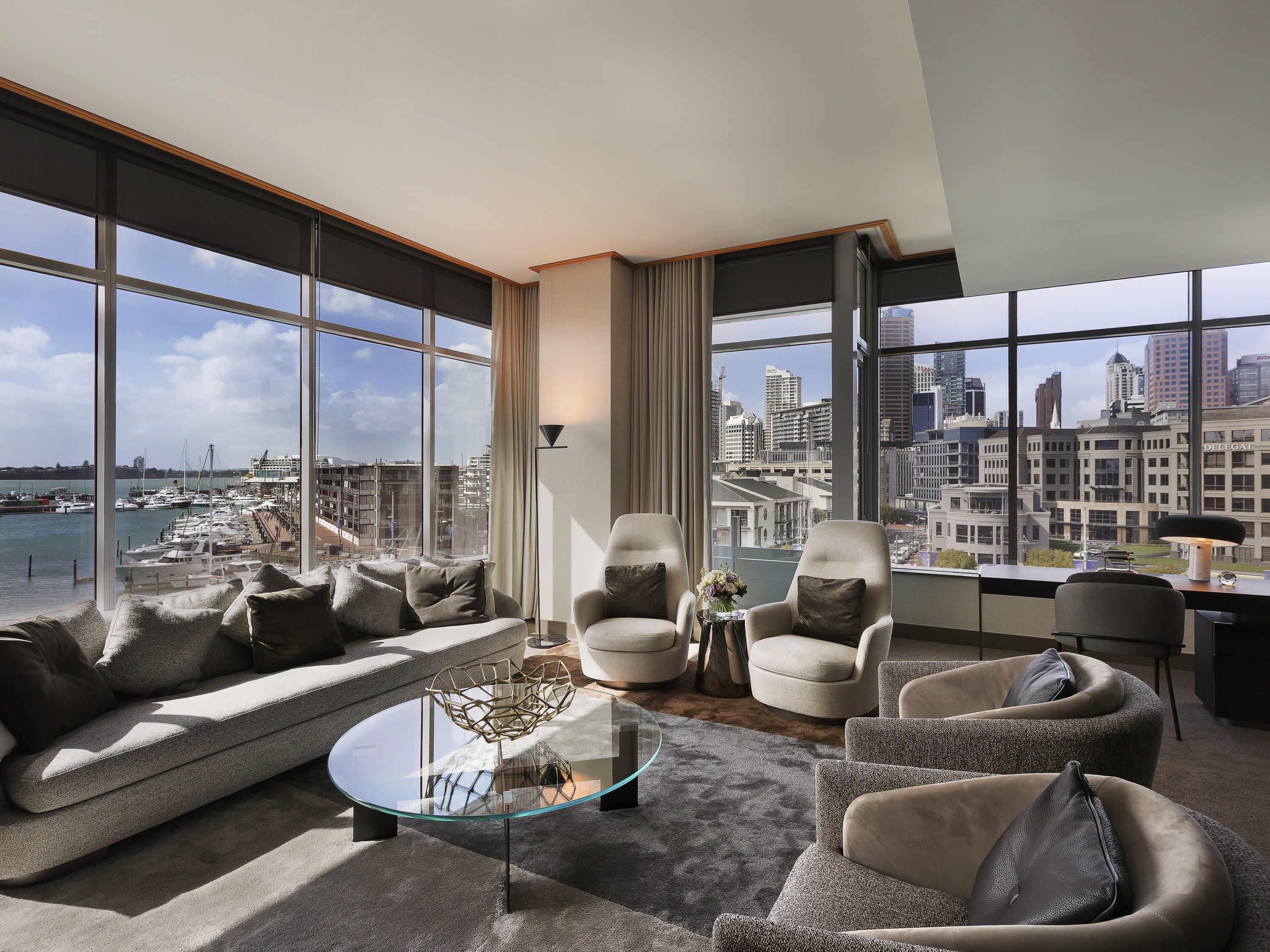 A modern living room with a sofa, armchairs, a glass coffee table, and a view of a marina and cityscape.