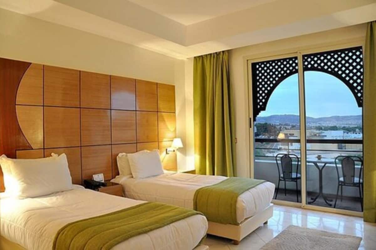 Habitación de hotel con dos camas, cabecera de madera, balcón con vistas y una mesa con sillas.