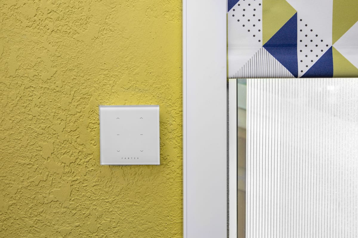 Un interruptor de luz en una pared amarilla con textura, una puerta blanca con un panel de vidrio con diseño geométrico y una pared blanca con textura.