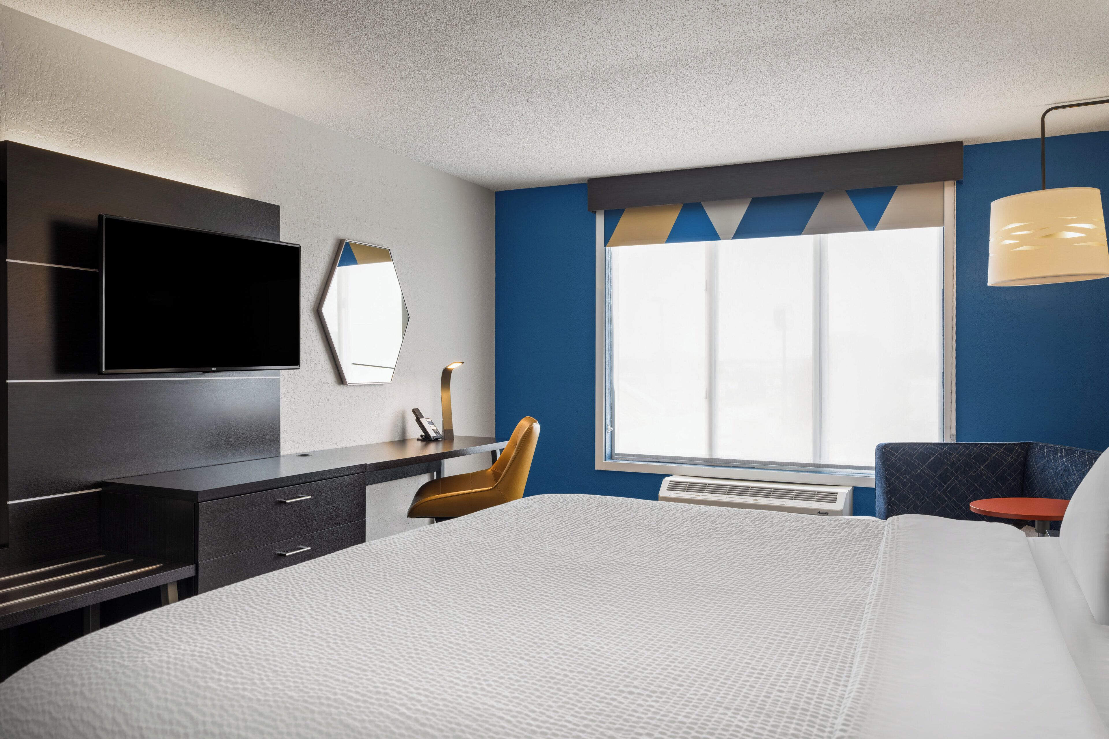 A hotel room with a bed, a desk, a chair, a television, a window, and a wall-mounted mirror.