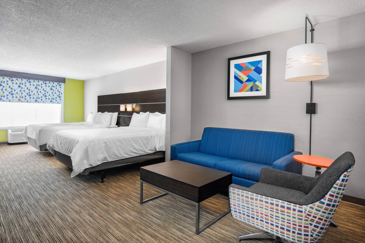 Habitación de hotel con dos camas, un sofá azul, un sillón estampado, una mesita naranja pequeña y una lámpara colgante moderna.