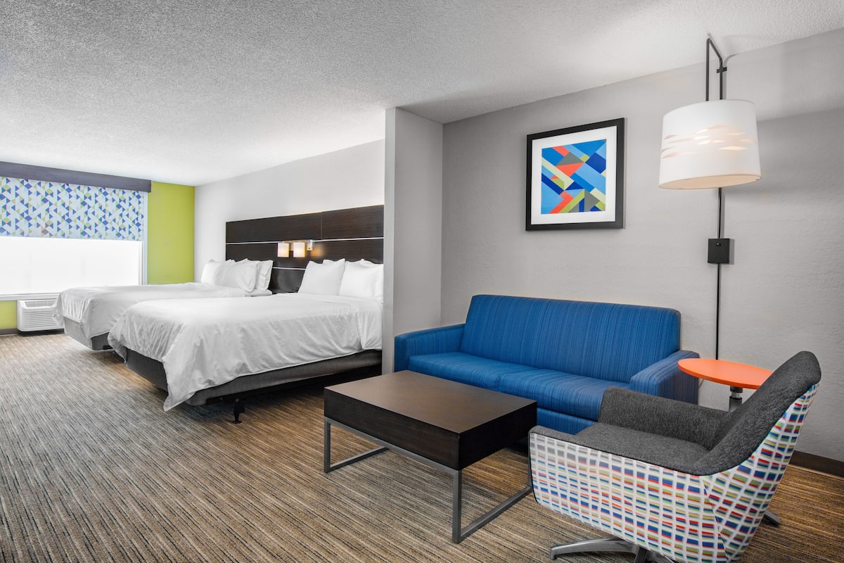 Habitación de hotel con dos camas, un sofá azul, un sillón estampado, una mesita naranja pequeña y una lámpara colgante moderna.