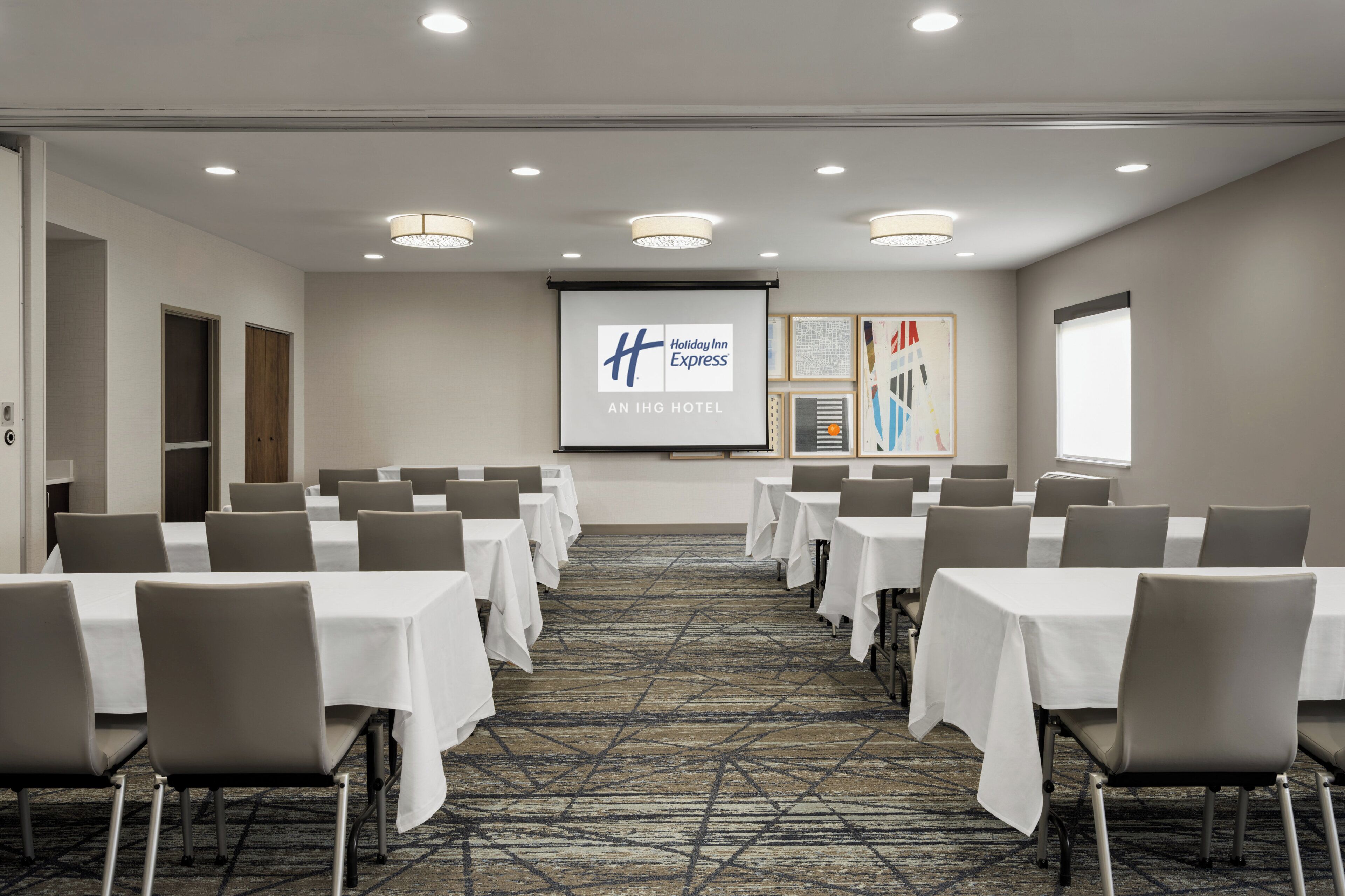 A conference room with rows of tables and chairs, a projector screen displaying the Holiday Inn Express logo, and framed artwork on the walls.