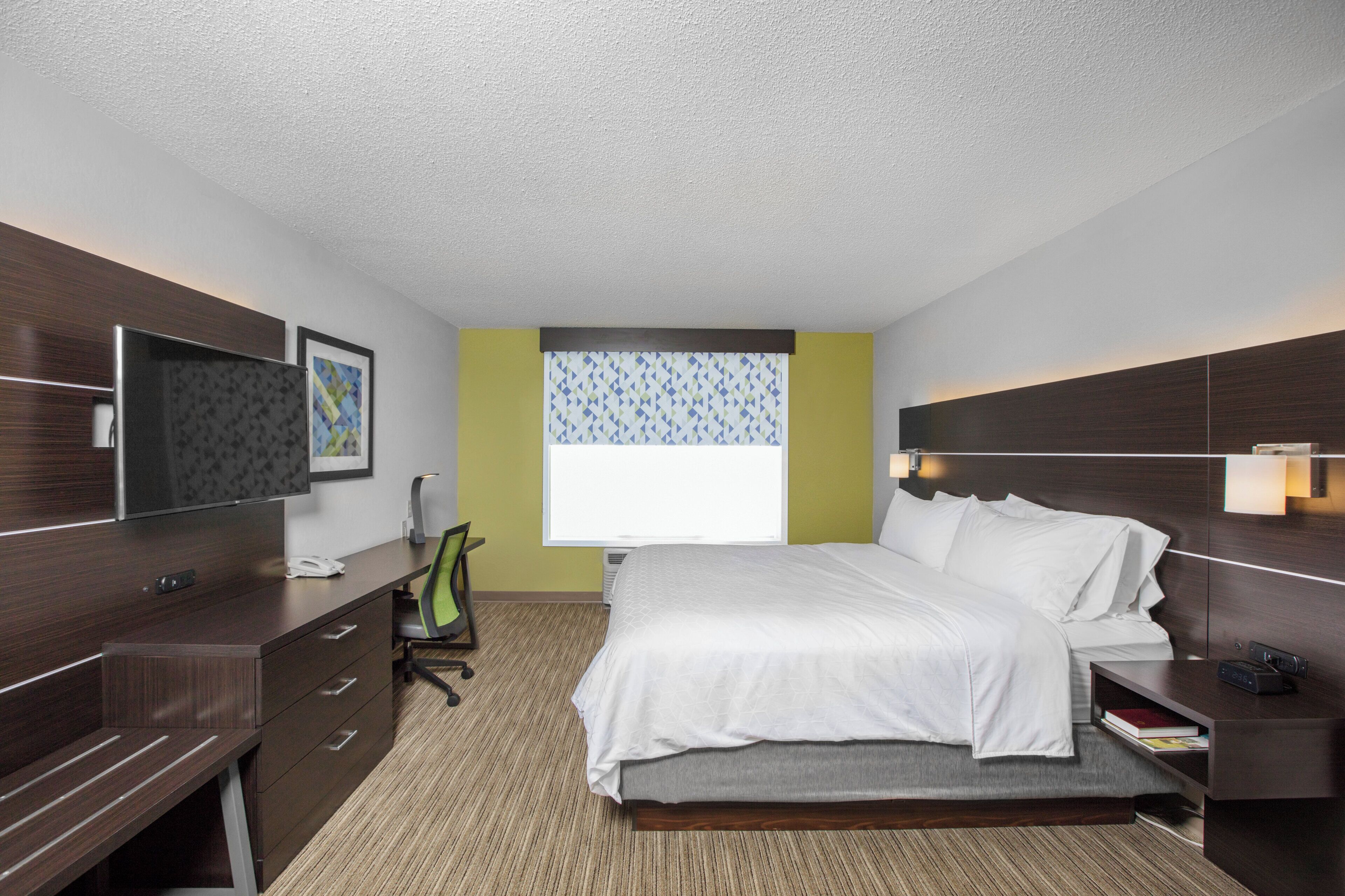 Habitación de hotel con cama, escritorio, silla, televisor y ventana con cortinas estampadas.