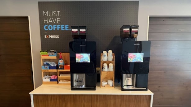 A coffee station with two espresso machines and a display of coffee cups and pods. - Holiday Inn Express Hotel & Suites Altoona-Des Moines by IHG (Altoona)