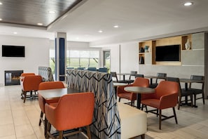 A modern dining area with orange chairs, a patterned sofa, and a flat-screen TV mounted on the wall. - Holiday Inn Express Hotel & Suites Altoona-Des Moines by IHG (Altoona)