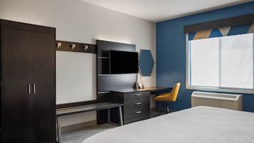 A hotel room with a bed, a desk with a chair, a wardrobe, a flat-screen TV, a mirror, and a window with an air conditioner.