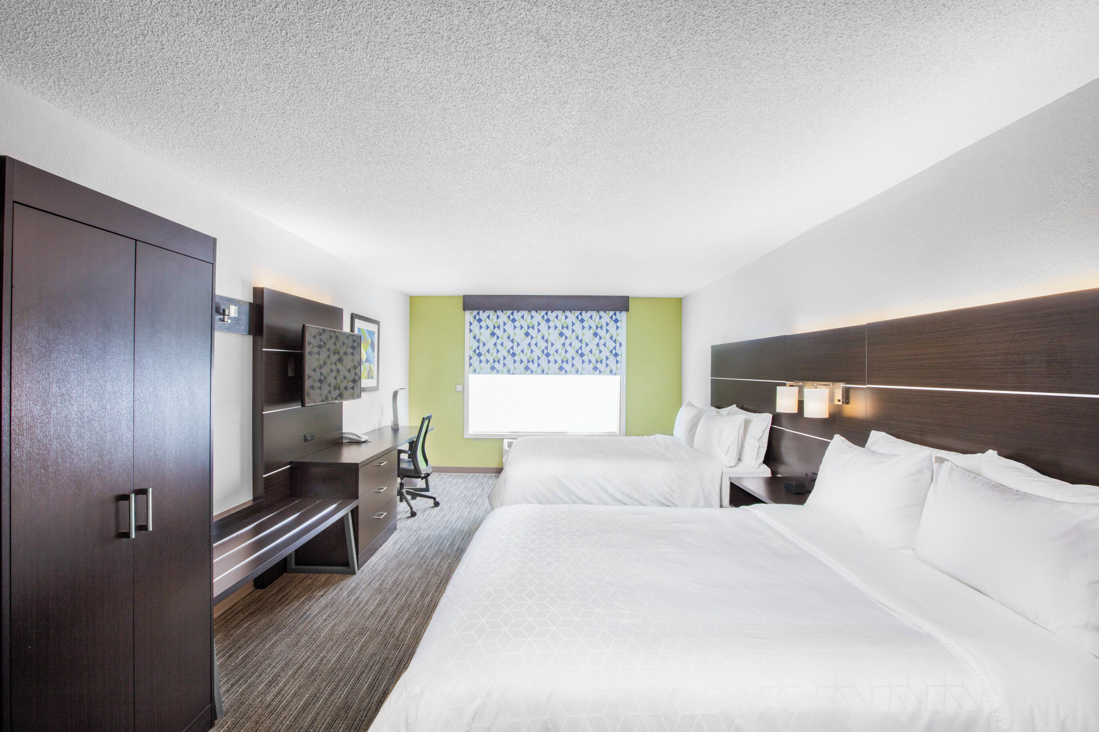 A hotel room with two beds, a desk, a chair, a TV, and a window with patterned curtains.