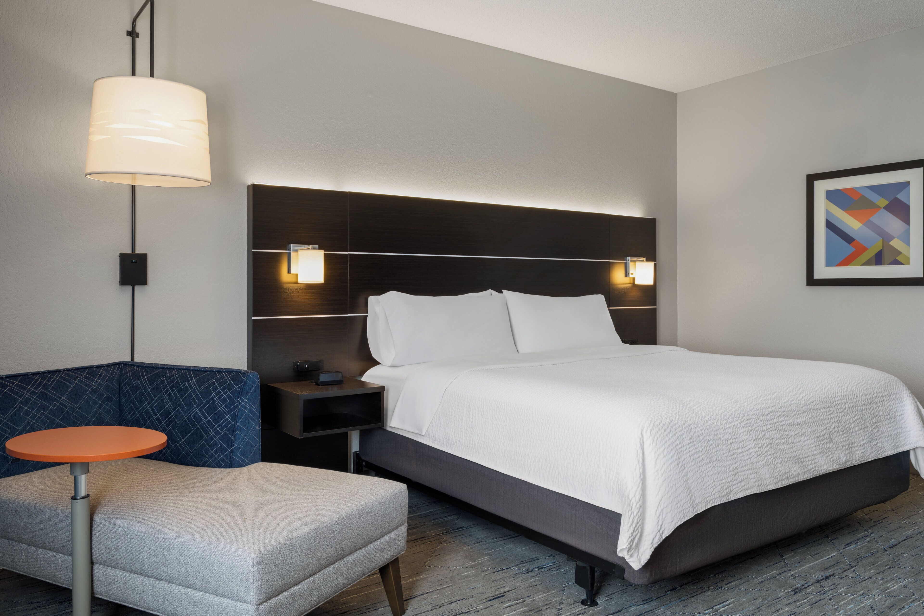 A hotel room with a bed, a nightstand, a lamp, a chair, and a framed picture on the wall.