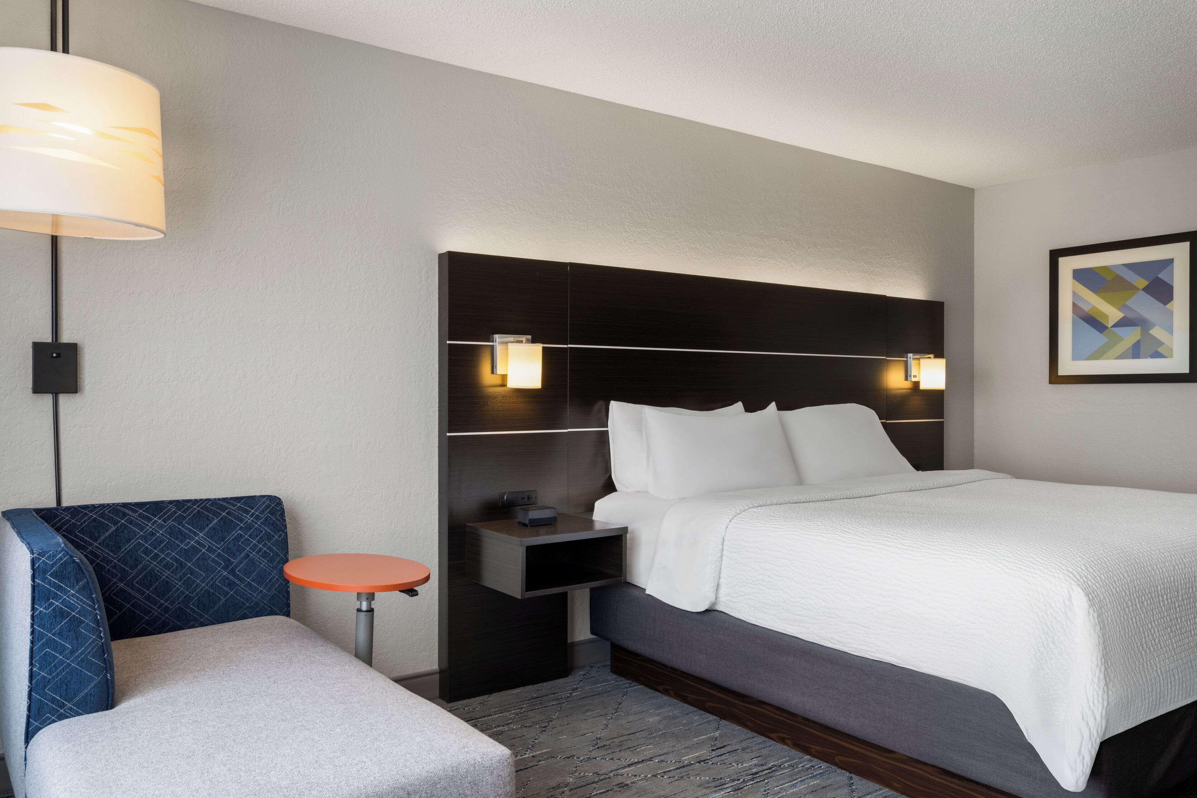 Quarto de hotel com cama, mesa de cabeceira, abajur e uma cadeira.