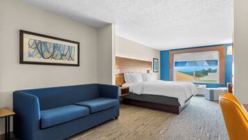 Habitación de hotel con sofá azul, cama con edredón blanco, cabecera de madera, ventana grande con vistas y un cuadro abstracto enmarcado en la pared.