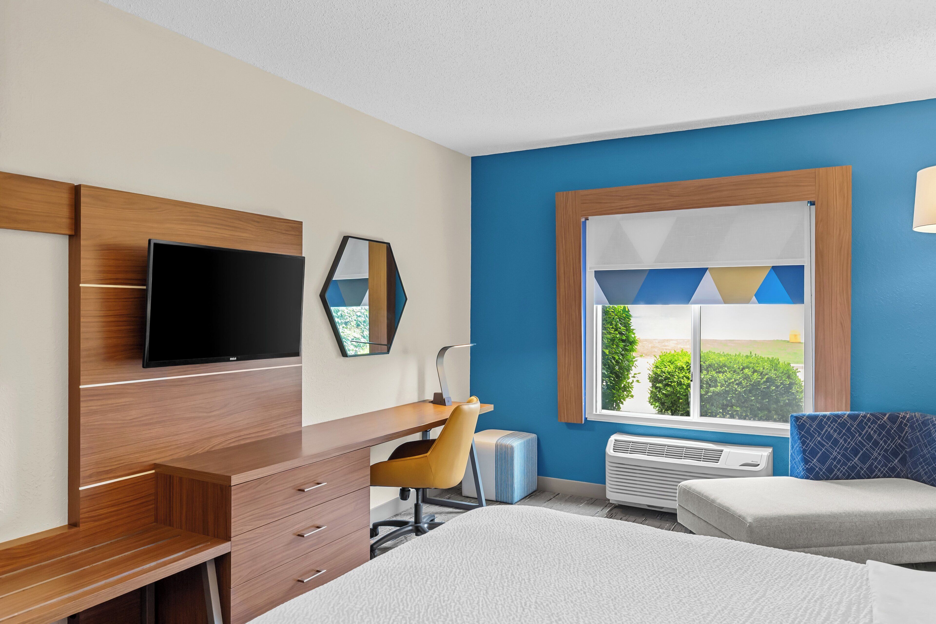 A hotel room with a bed, a desk with a chair, a flat-screen TV, a window with a view, and an air conditioning unit.