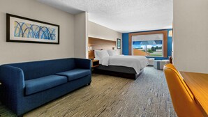 A hotel room with a blue sofa, a bed with white bedding, a nightstand, a desk with a chair, and a large window offering a view. - Holiday Inn Express & Suites Vandalia by IHG (Vandalia)