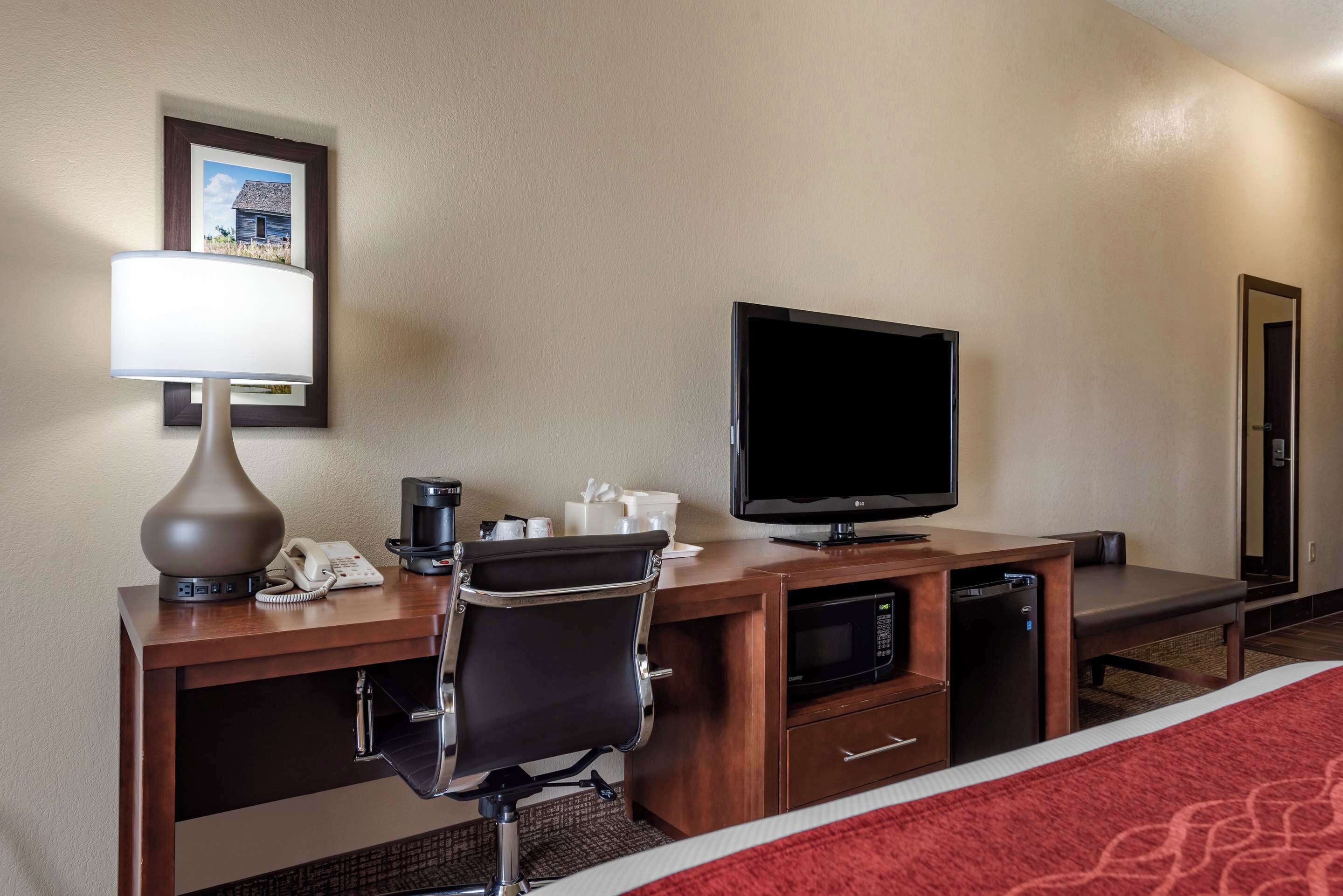 Habitación de hotel con escritorio de madera, televisor de pantalla plana, lámpara, teléfono, microondas y cama con manta roja.
