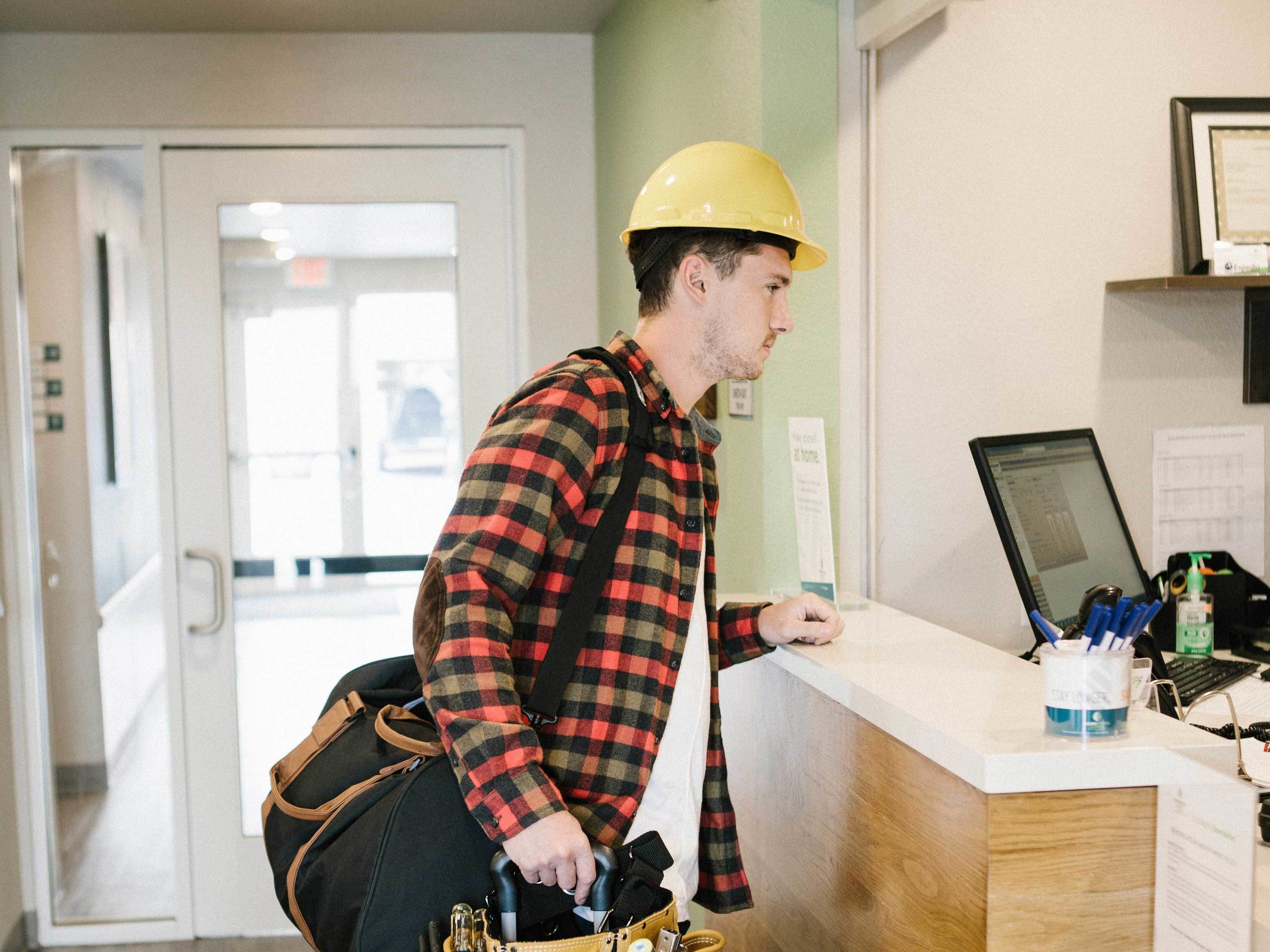 Eine Person in kariertem Hemd und gelbem Schutzhelm steht an einer Rezeption mit einem Laptop, Werkzeugen und einer Tasche.