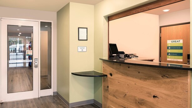 A reception area with a wooden reception desk, a glass partition, and a signboard. - Extended Stay America Select Suites - Fayetteville (Fayetteville)