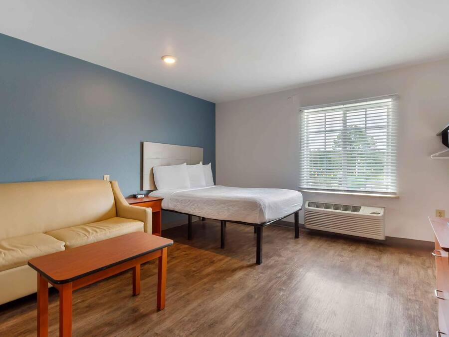 Quarto com cama, sofá, mesa pequena, escrivaninha com computador e janela com persianas.