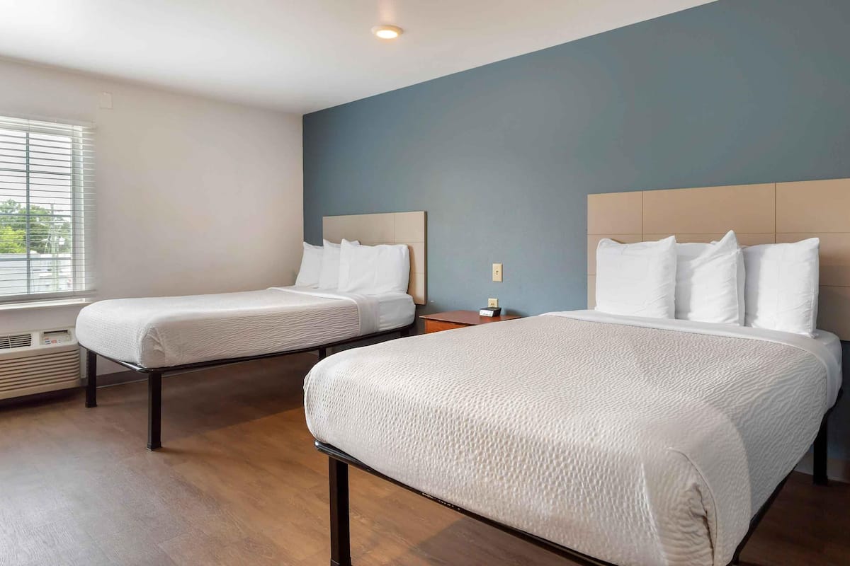 A hotel room with two beds, a window with blinds, a bedside table with a lamp, and a wall heater.