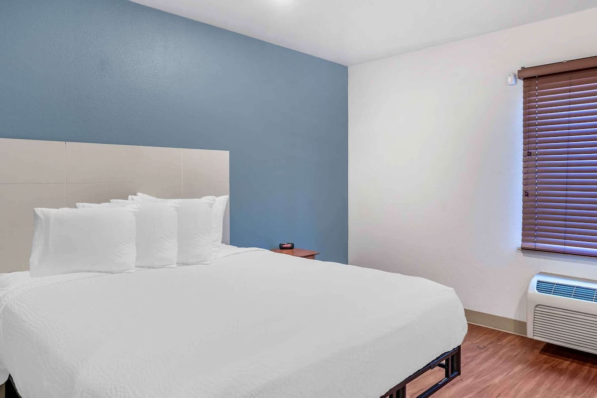 A neatly made bed with white bedding in a room with a blue accent wall and a window with blinds.