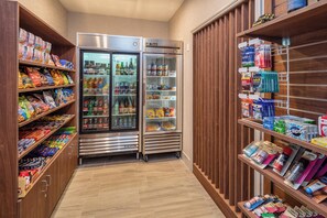 A narrow hallway with a refrigerator stocked with beverages and snacks, adjacent to shelves filled with various food items and toiletries.