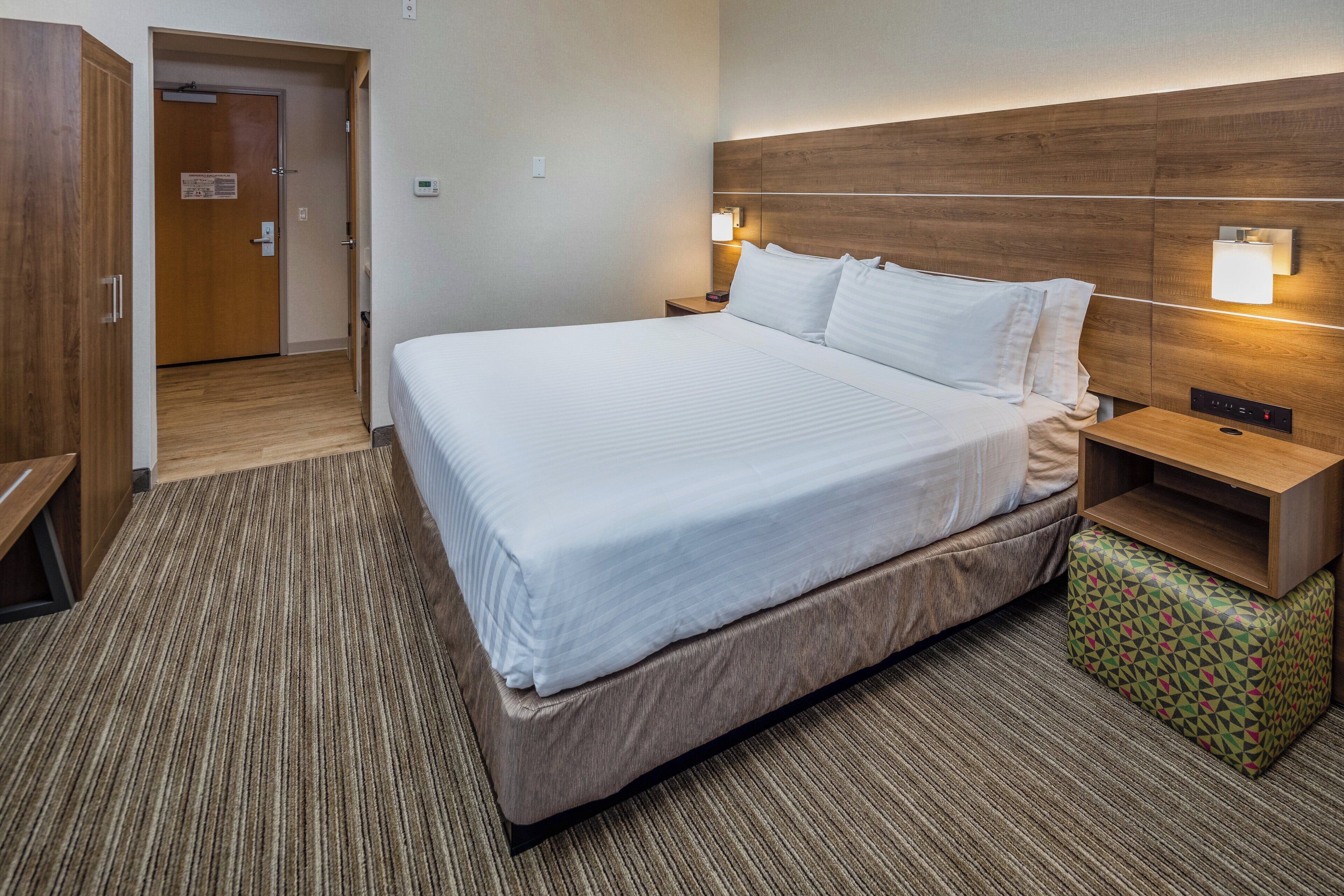 A hotel room with a large bed, wooden headboard, bedside lamps, a small wooden nightstand, and a patterned ottoman.
