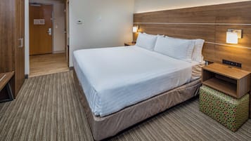 A hotel room with a large bed, wooden headboard, bedside lamps, a small wooden nightstand, and a patterned ottoman.