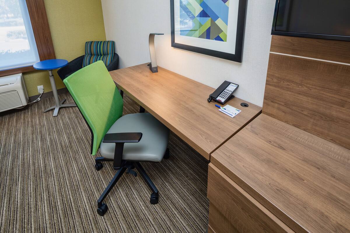 A hotel room with a wooden desk, a green office chair, a blue chair, a framed picture on the wall, and a window with a view of snow outside.