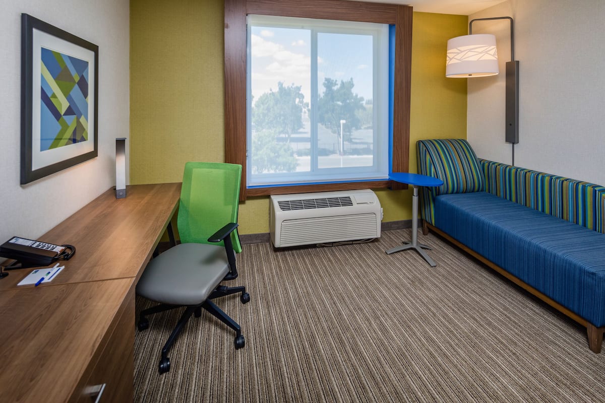 A hotel room with a desk, chair, air conditioning unit, sofa, and a window with a view of trees.