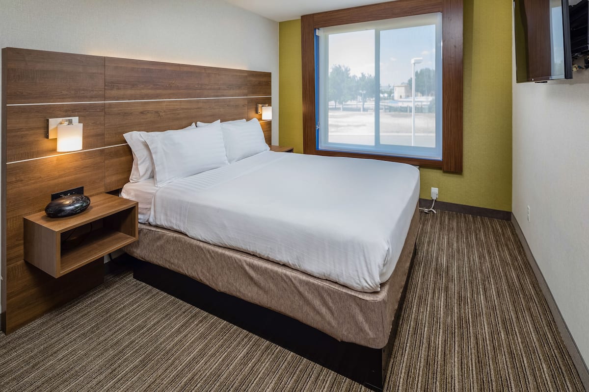 A hotel room with a large bed, wooden headboard, bedside lamps, a small shelf, and a window with a view of the outside.
