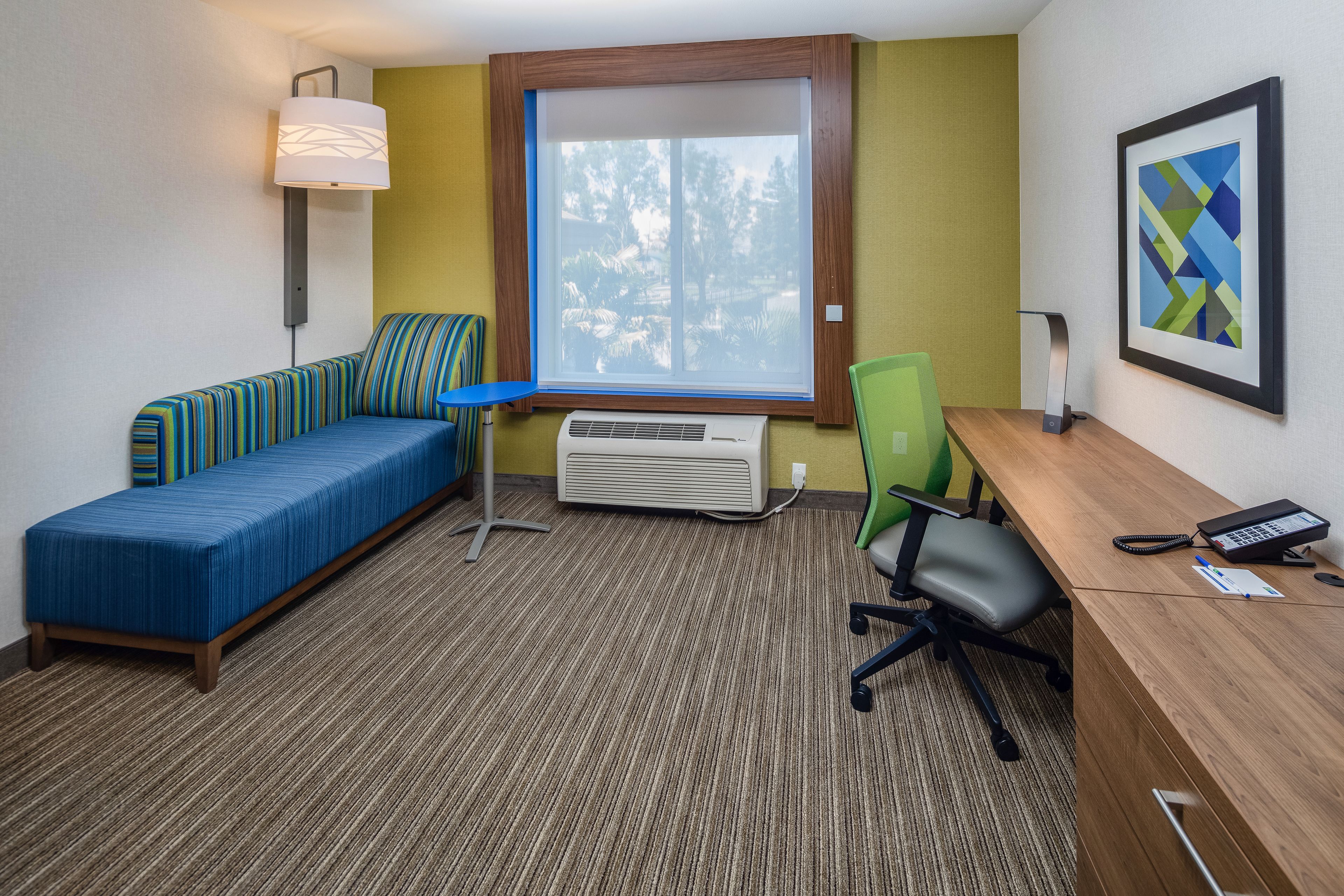 Ein Hotelzimmer mit blauem Sofa, einem Schreibtisch mit Stuhl, einem Fenster mit Blick auf Bäume, einer Klimaanlage und einem gerahmten Bild an der Wand.
