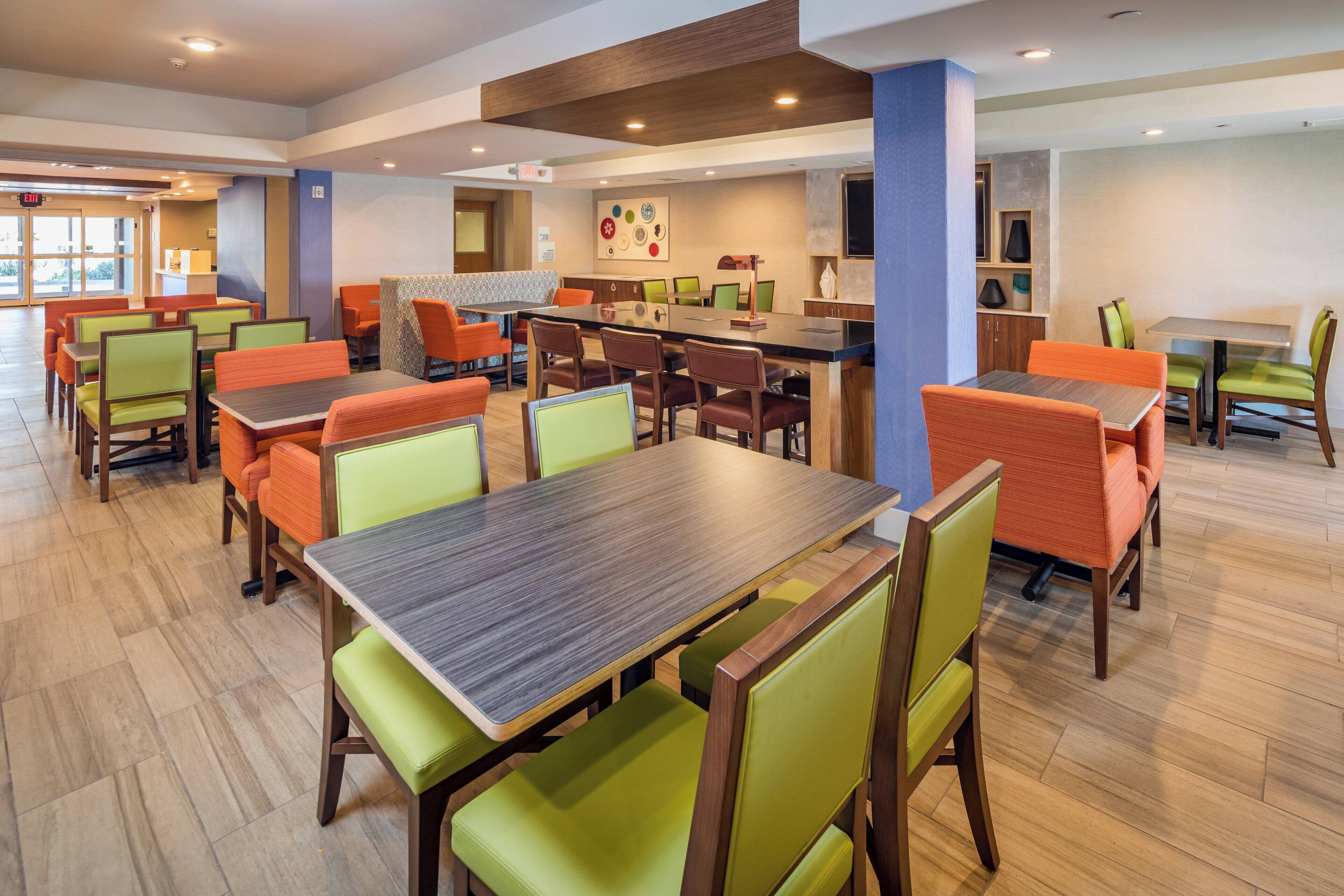 A hotel dining area with multiple tables and chairs, a central bar area, and a view of the outdoors through large windows.