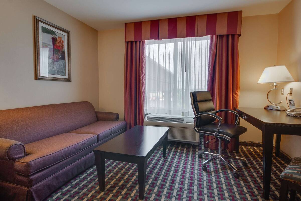 A hotel room with a sofa, a small table, a chair, a desk with a lamp, and a window with curtains.