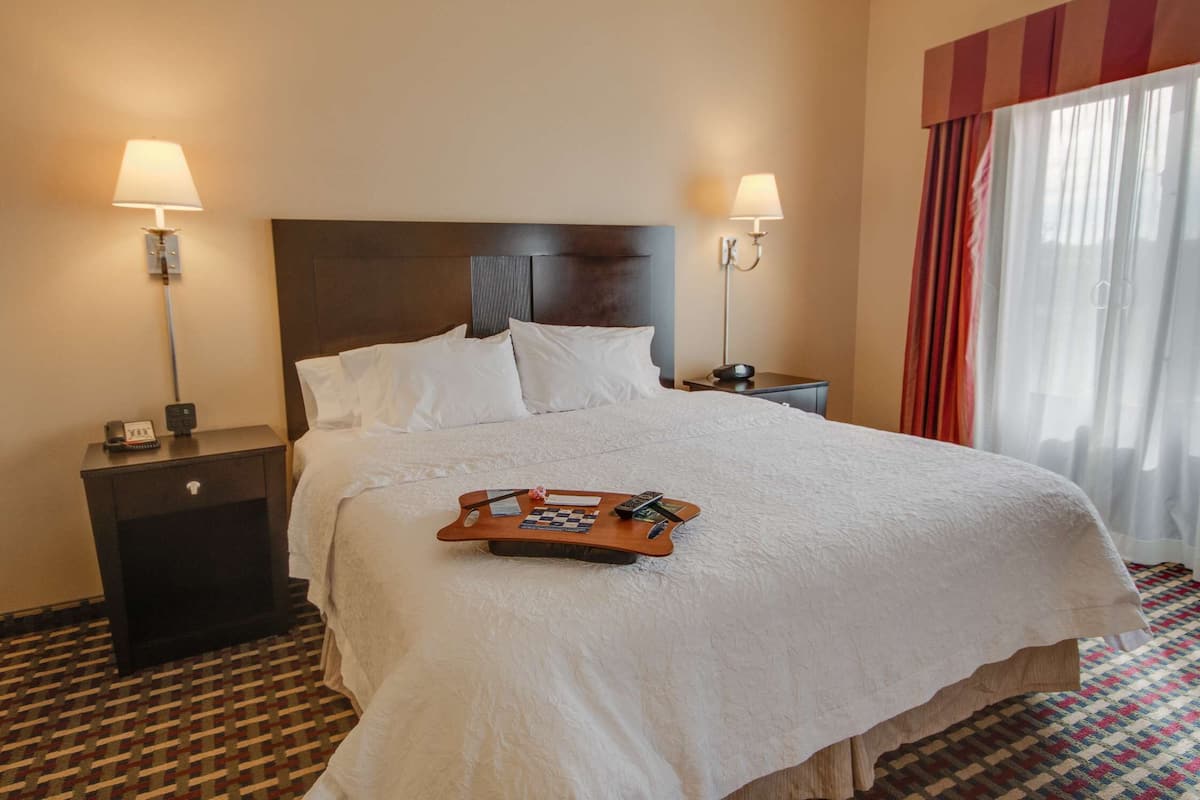 A hotel room with a large bed, two bedside tables, a nightstand, a tray table, and a window with curtains.