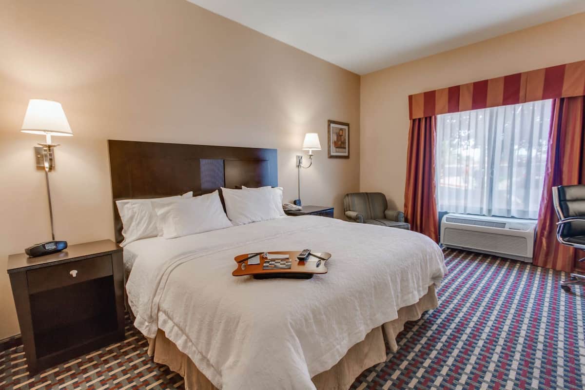A hotel room with a large bed, two bedside lamps, a chair, a window with curtains, and a tray on the bed.