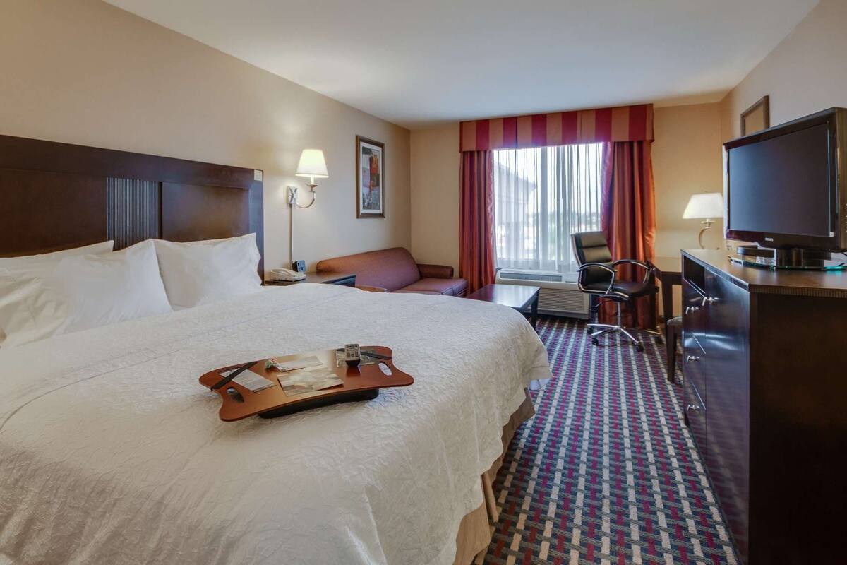A hotel room with a large bed, a desk with a television, a chair, and a desk lamp.