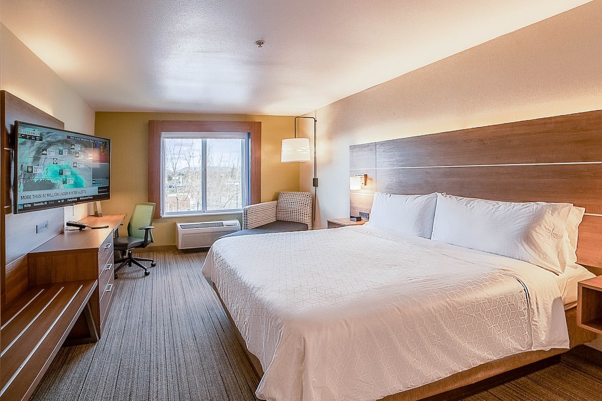 A hotel room with a large bed, a desk with a computer, a chair, a window, and a TV displaying weather information.