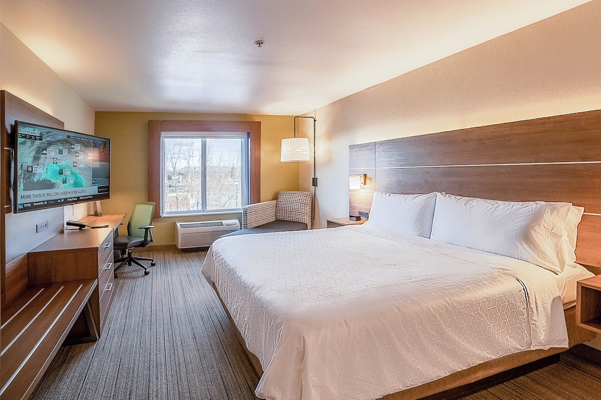 A hotel room with a large bed, a desk with a computer, a chair, a window, and a TV displaying weather information.