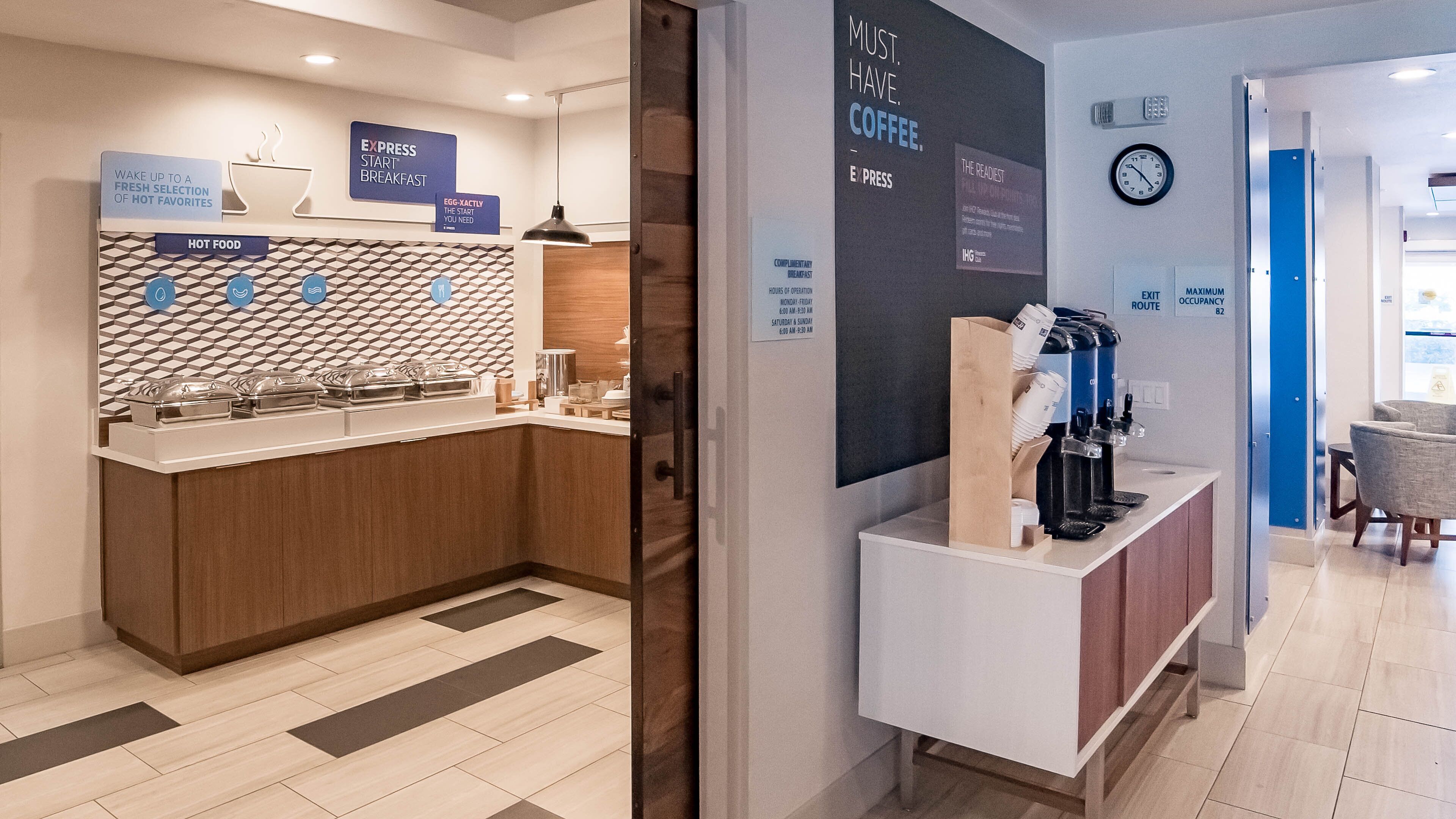 A modern hotel breakfast area with a buffet counter, coffee station, and a sign indicating the availability of complimentary breakfast.