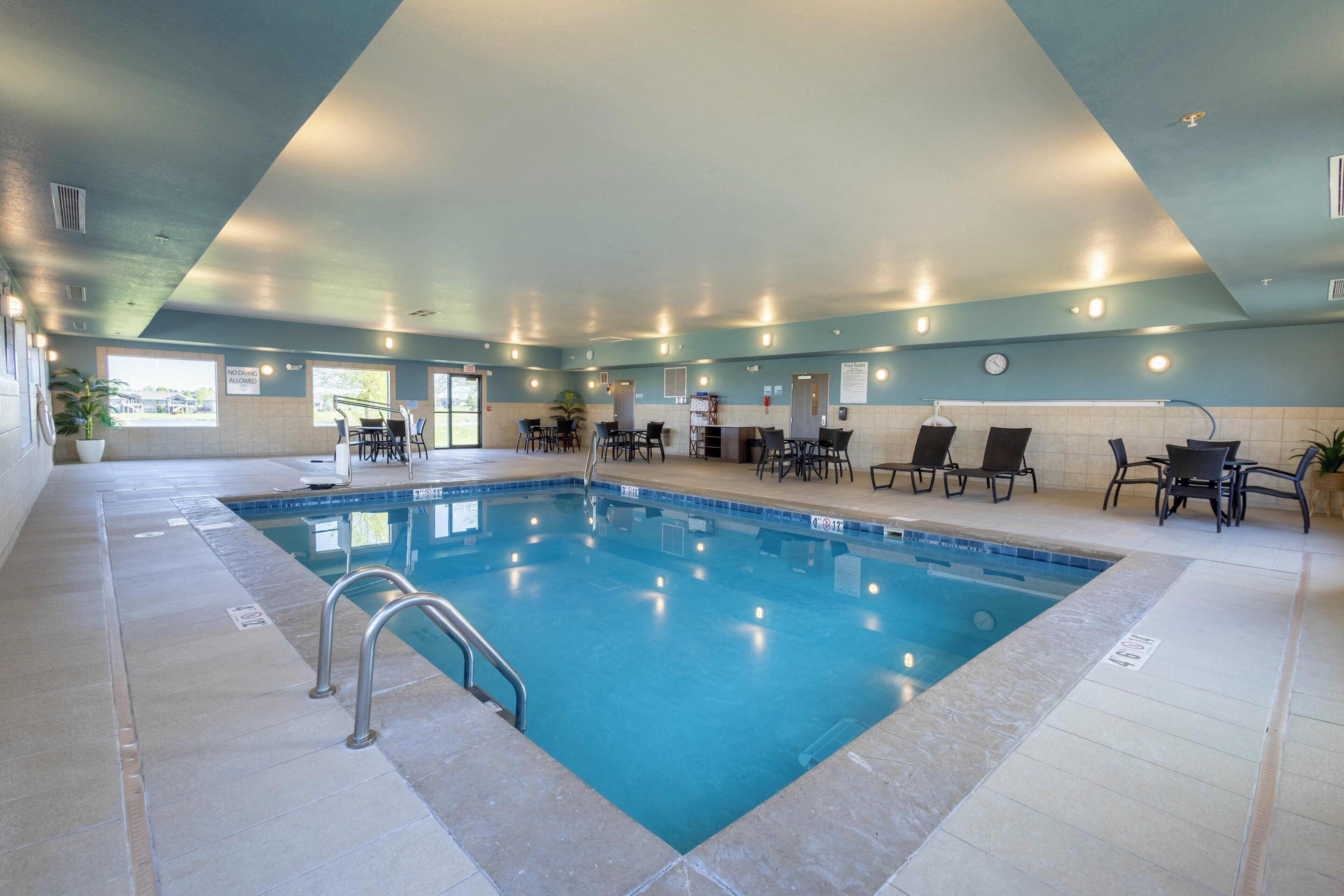 An indoor swimming pool with a tiled floor, a metal ladder, and several chairs around the pool area.