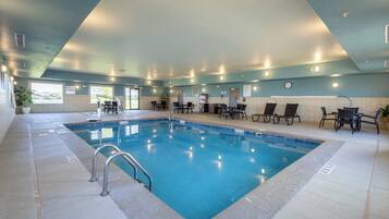 An indoor swimming pool with a tiled floor, a metal ladder, and several chairs around the pool area.