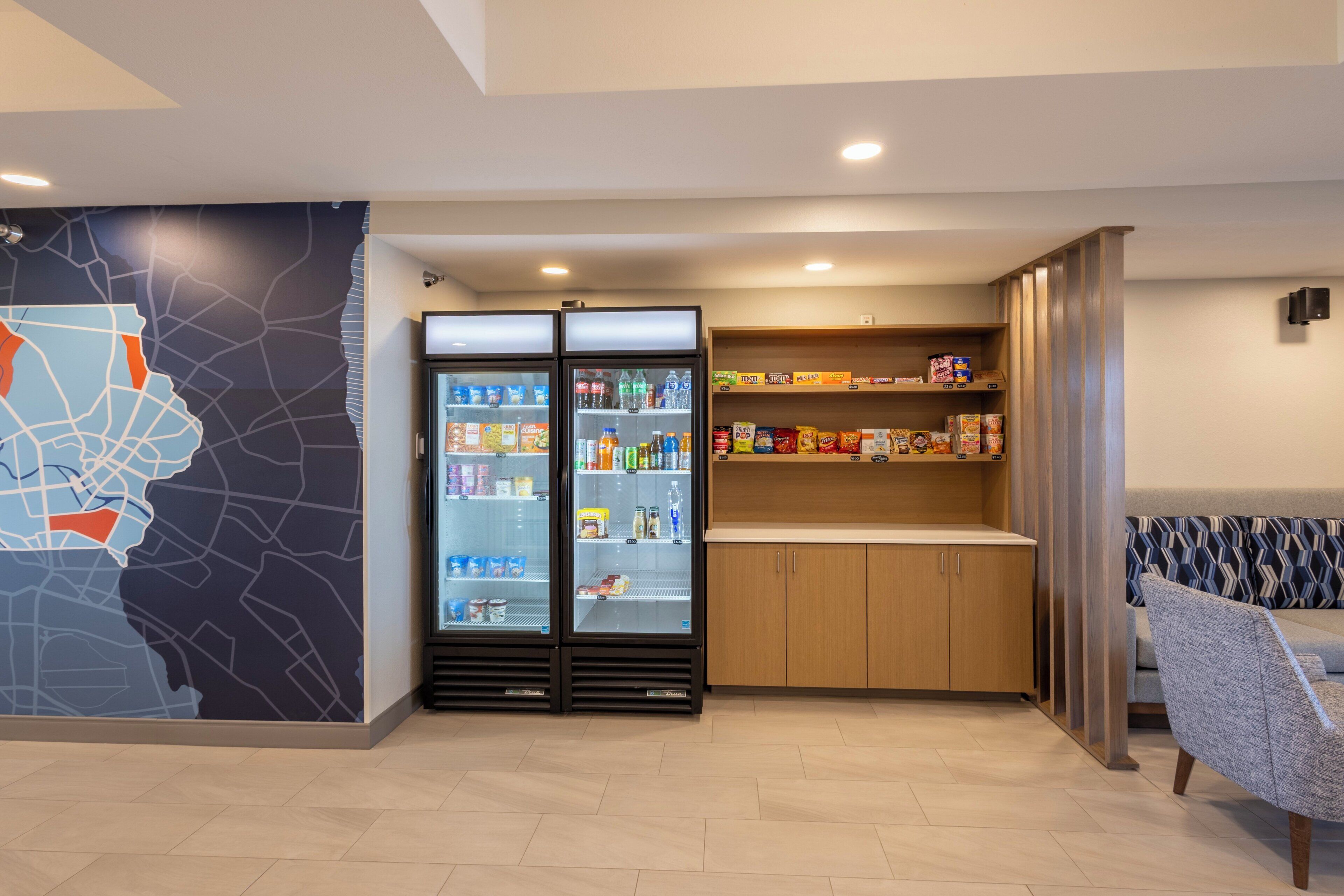 A modern hotel lobby with a map mural, a refrigerated beverage display, a snack bar, and a seating area.