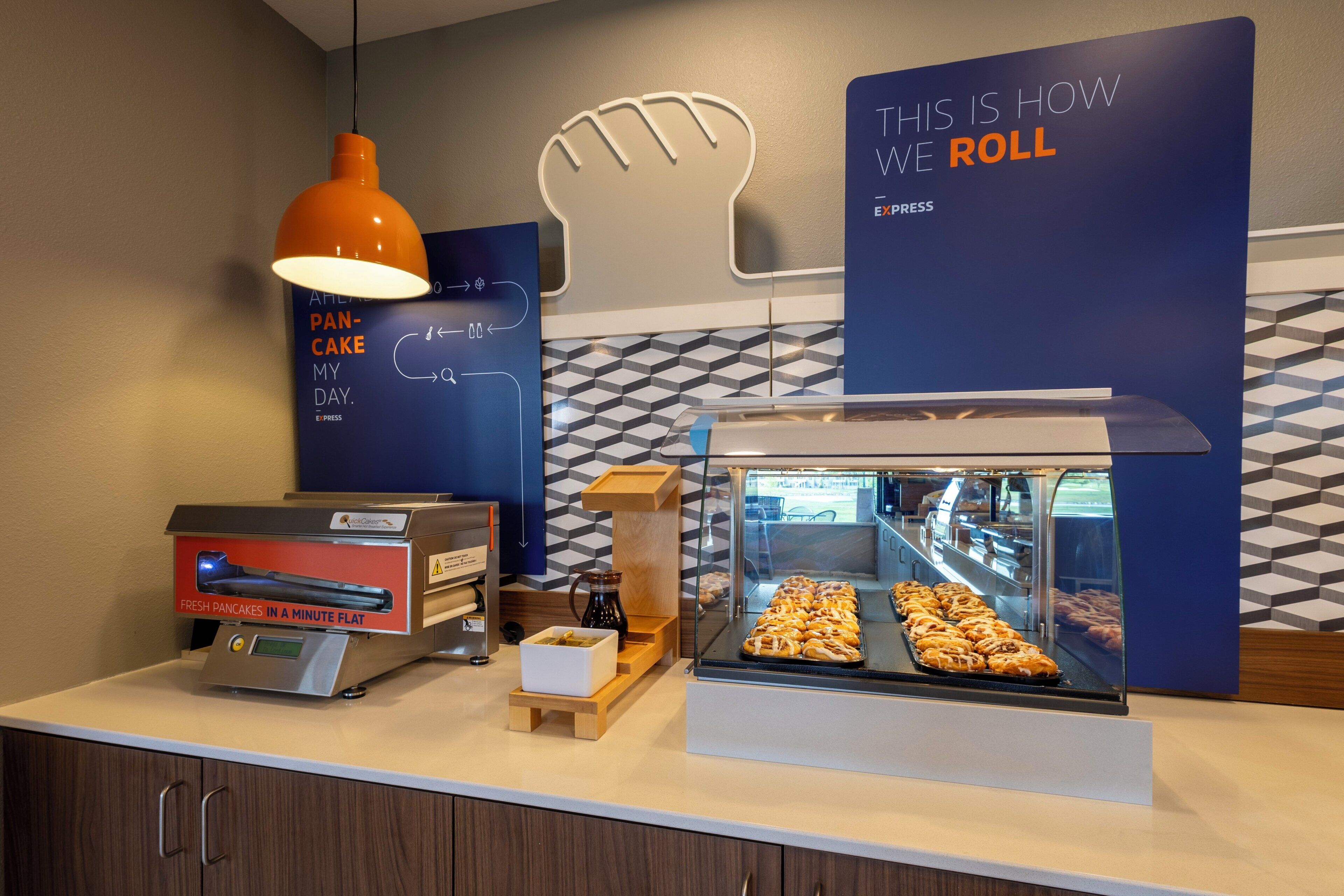 A breakfast counter with a coffee machine, a display case with pastries, and a wall with a geometric pattern and a slogan sign.